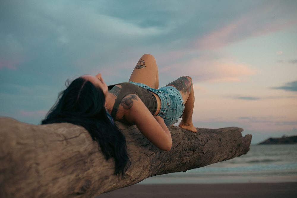 A tattooed woman relaxes on a driftwood log at a beach during a colorful sunset.