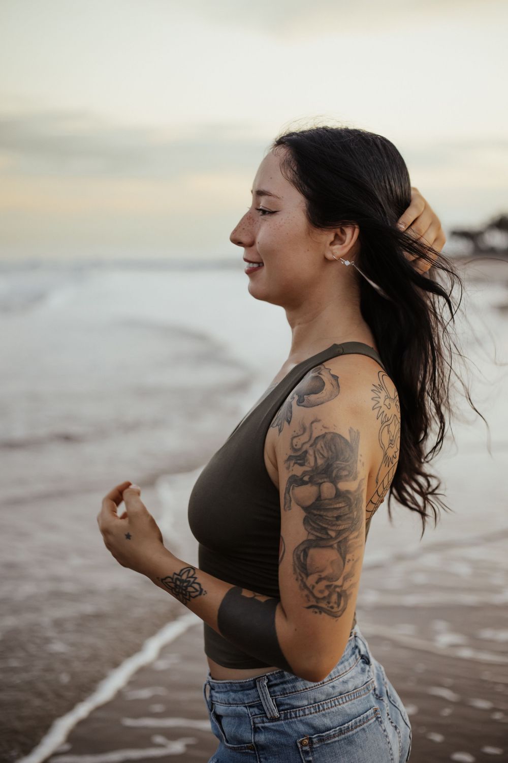 Smiling tattooed woman in olive crop top and jeans standing on beach at sunset with wind in her hair.
