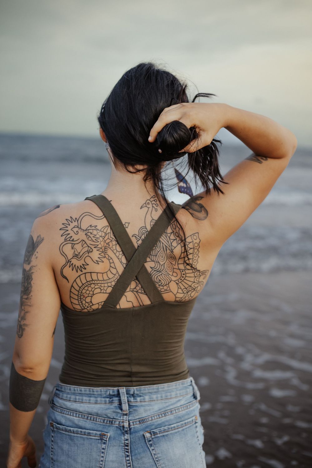 Woman with dragon tattoos on her back and arm stands facing the ocean wearing a green top and denim shorts.