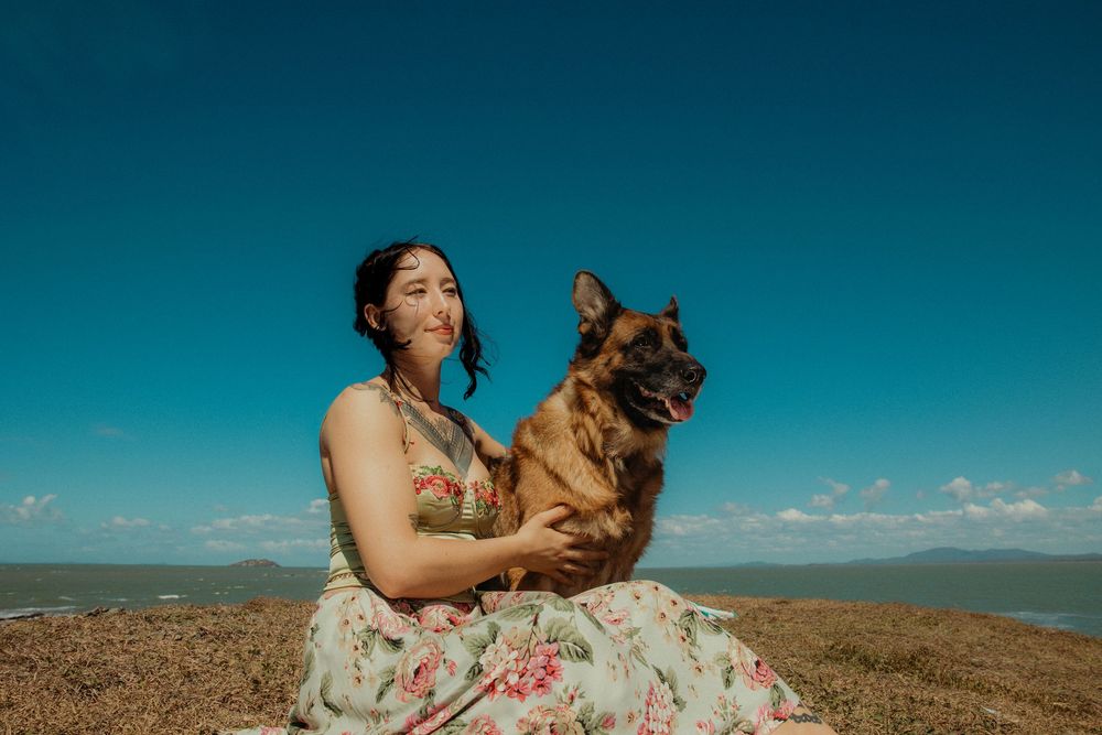 Woman in floral dress sitting outdoors holding a German Shepherd dog with blue sky and ocean in background.