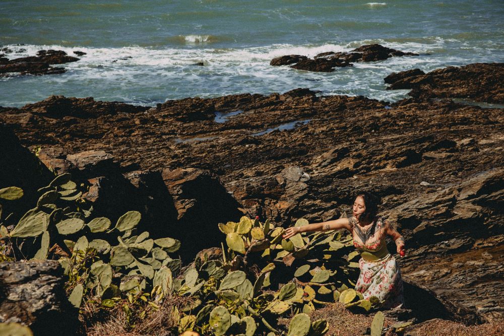 Two people sitting among cactus plants on rocky coastal cliffs with ocean waves in the background.