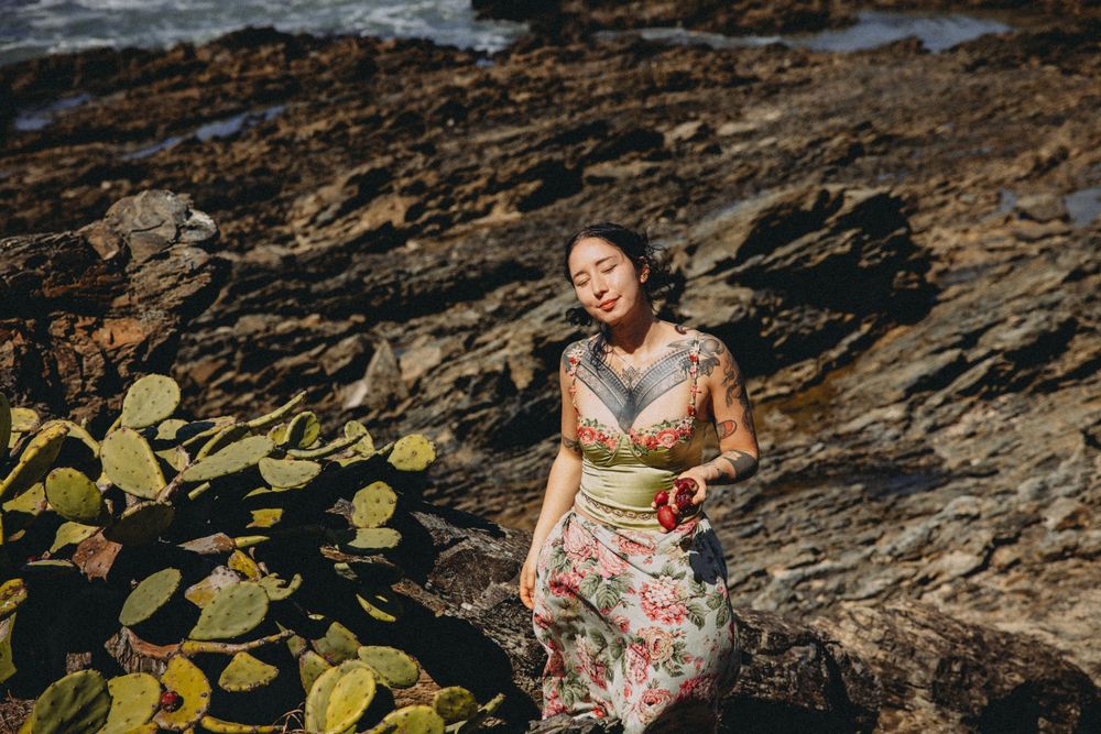 Tattooed woman in floral dress poses among coastal rocks and succulents in a serene outdoor portrait.