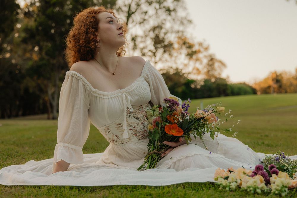 Woman with curly auburn hair in a white peasant-style dress sits on a green lawn holding a colorful bouquet of wildflowers.