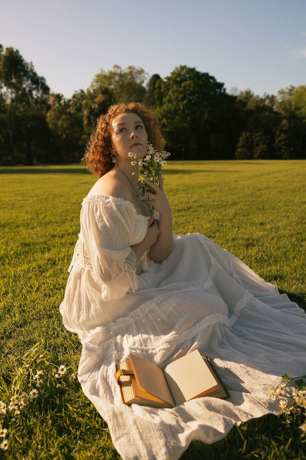 Woman with curly red hair in a white off-shoulder dress sits on a sunlit grass field holding wildflowers beside an open book.