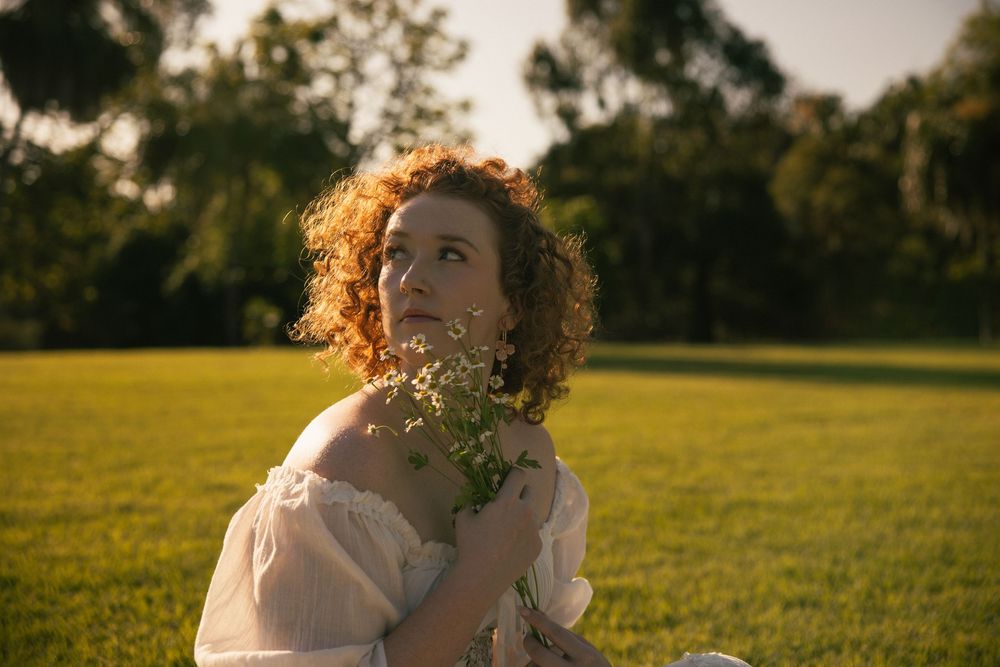 Woman with curly auburn hair in a white off-the-shoulder dress holds a bouquet of wildflowers while gazing upward on a sunlit grassy field.