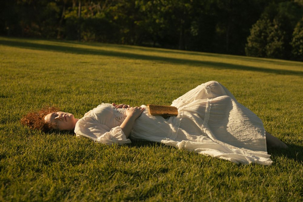Red-haired woman in a flowing white dress lying on a sunlit green lawn, holding a book, with tall trees in the background.
