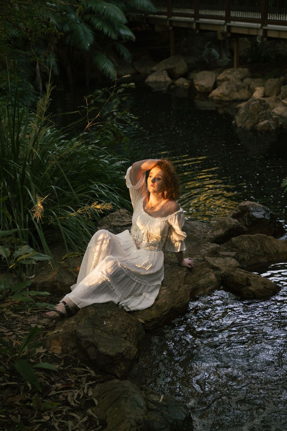 Woman in a flowing white dress sits on mossy rocks beside a dark pond, one hand raised to her head, surrounded by lush green plants.