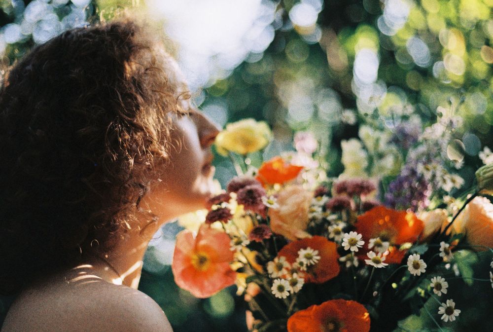 Woman with curly dark hair in profile smelling a vibrant bouquet of orange poppies, yellow blooms and white wildflowers in soft sunlight.