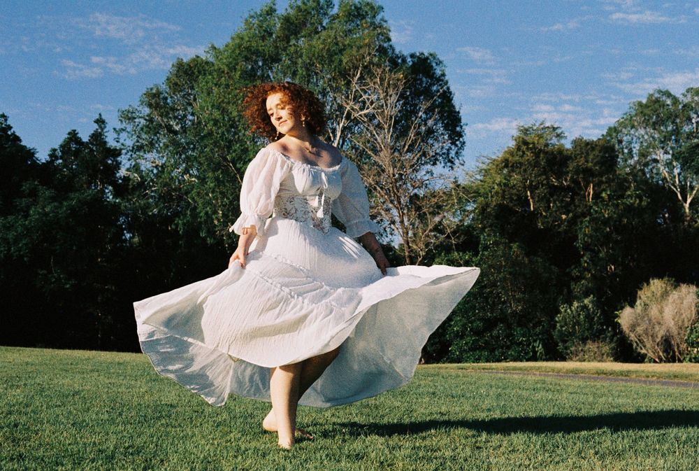 Woman in a flowing white dress twirls barefoot on a sunlit green lawn beneath tall trees and a blue sky.