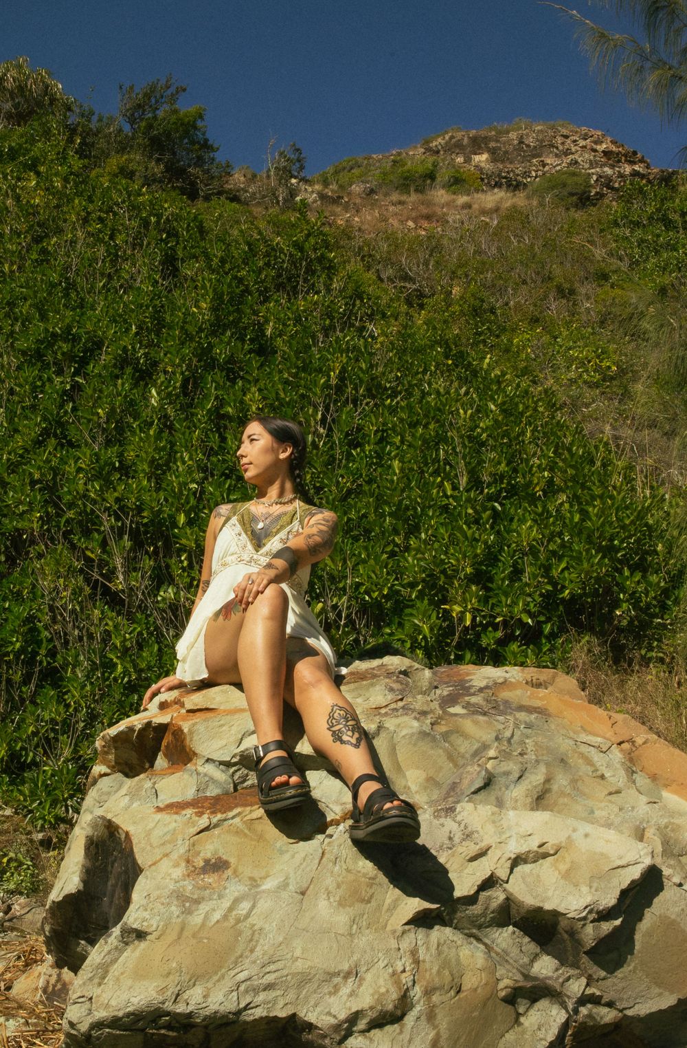 Tattooed woman in white dress and sandals sitting on a rock surrounded by lush green hillside vegetation.