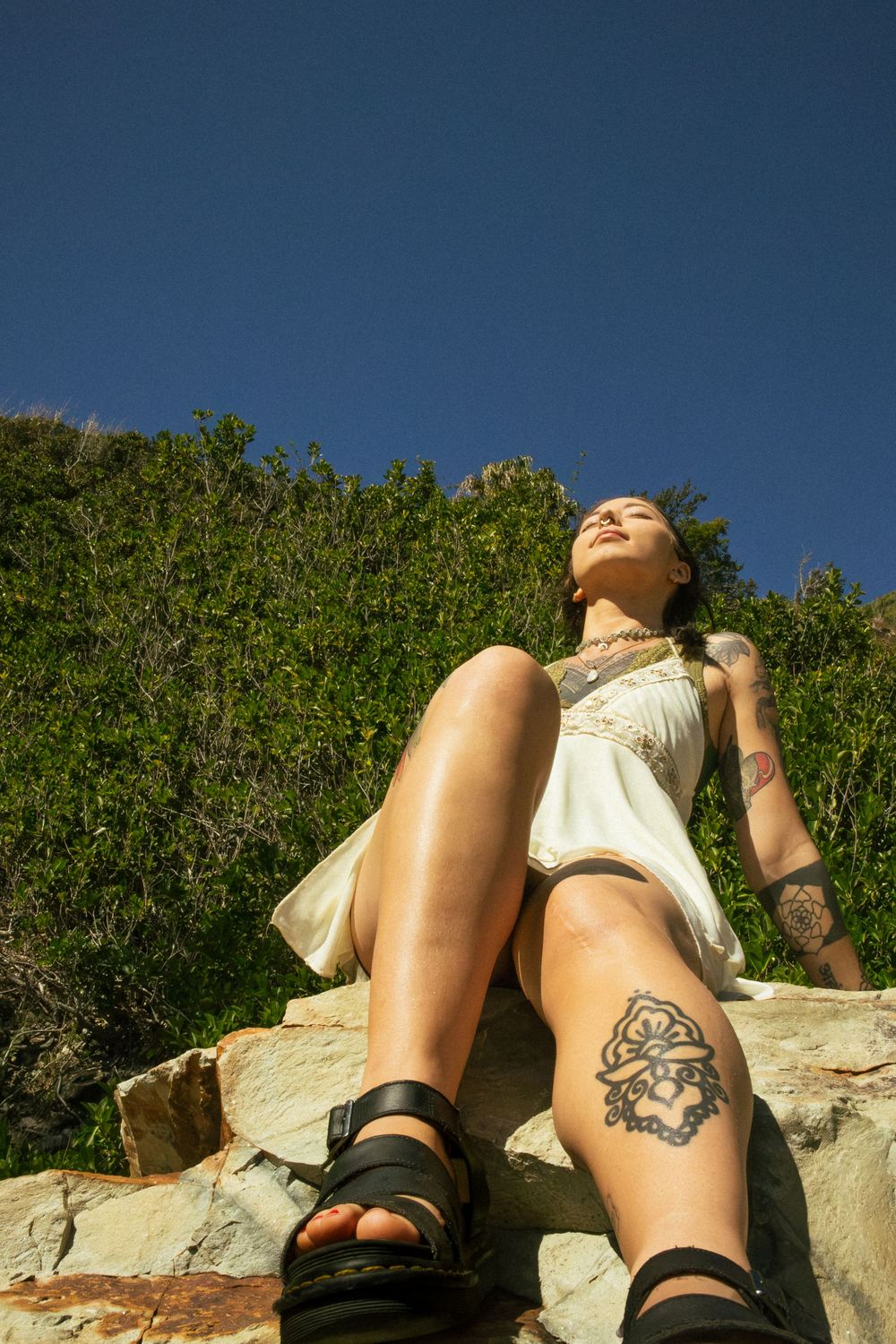 Woman with tattoos in white dress sitting on rocks outdoors, enjoying sunshine against blue sky and green hillside.