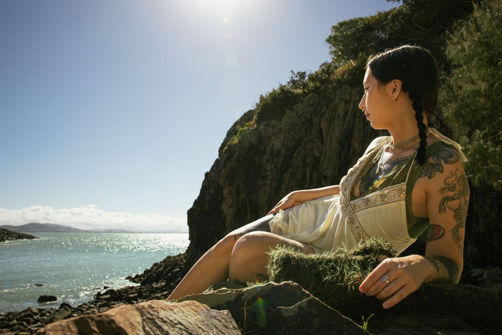 Tattooed woman in white dress sitting on coastal rocks overlooking turquoise ocean under bright sunny sky.
