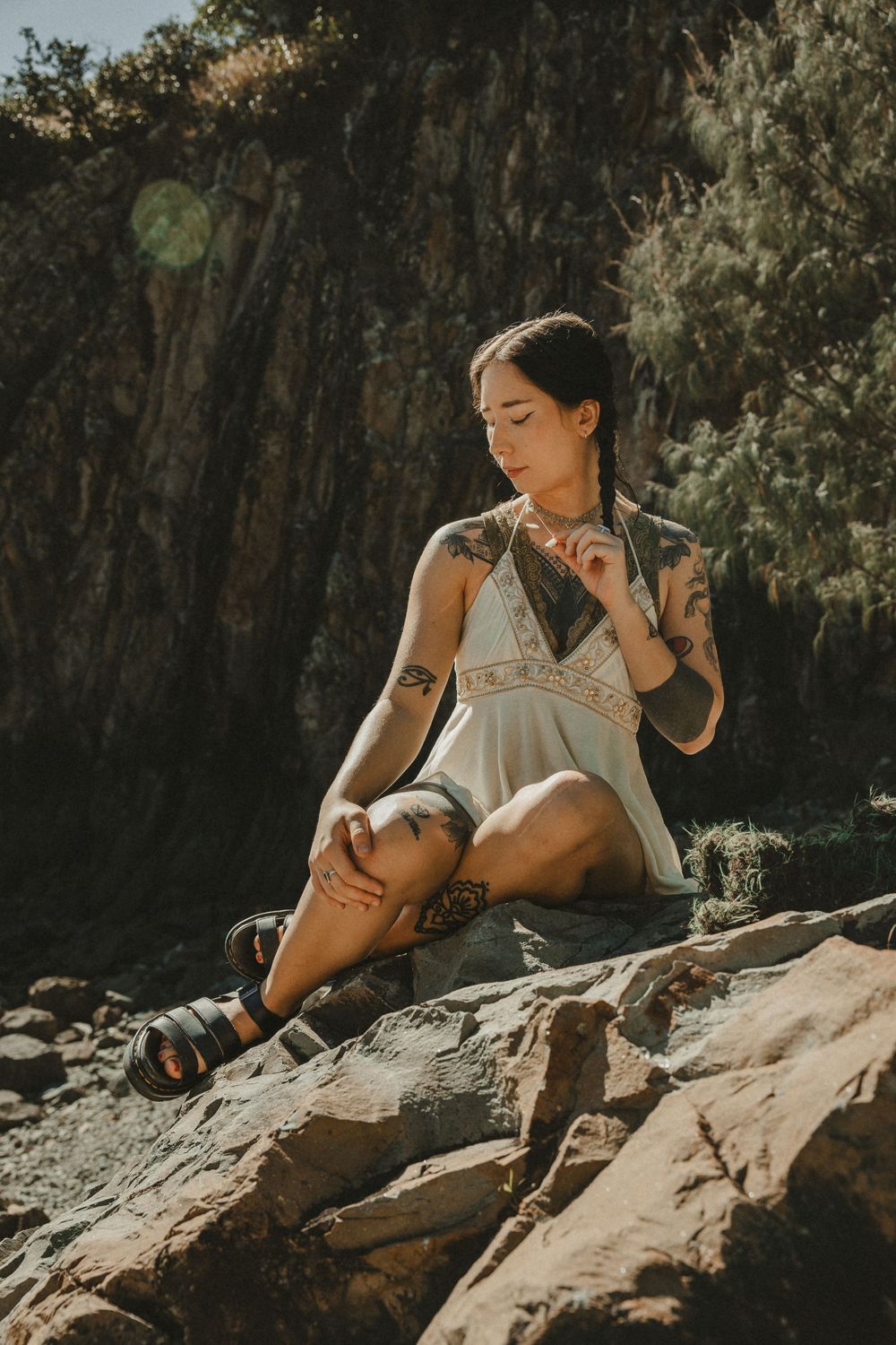 Tattooed woman in white dress and sandals sitting on rocks outdoors in natural sunlight with rocky cliffs behind her.