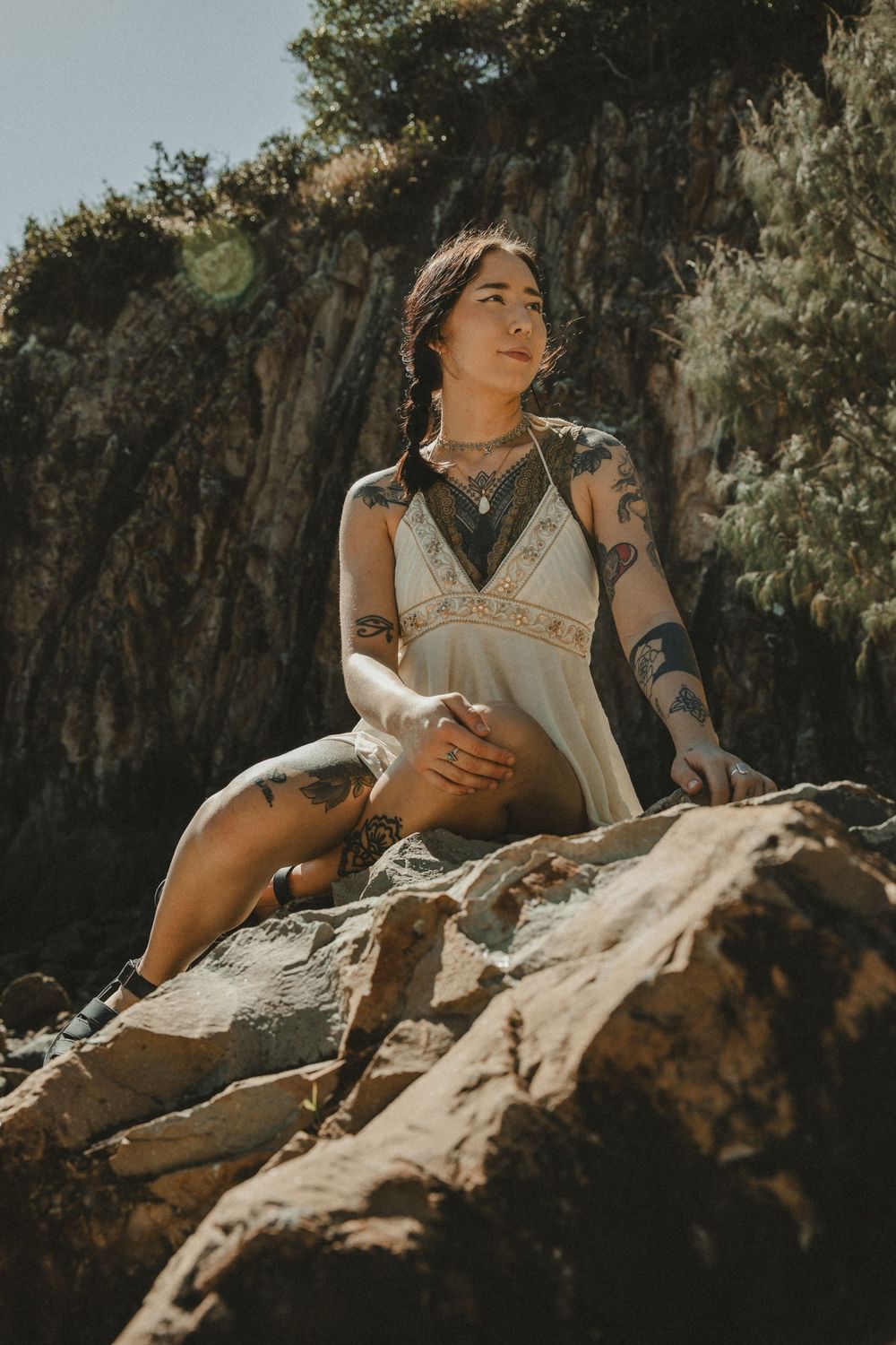 Tattooed woman in white dress sitting on rocks outdoors, gazing away in natural sunlight.