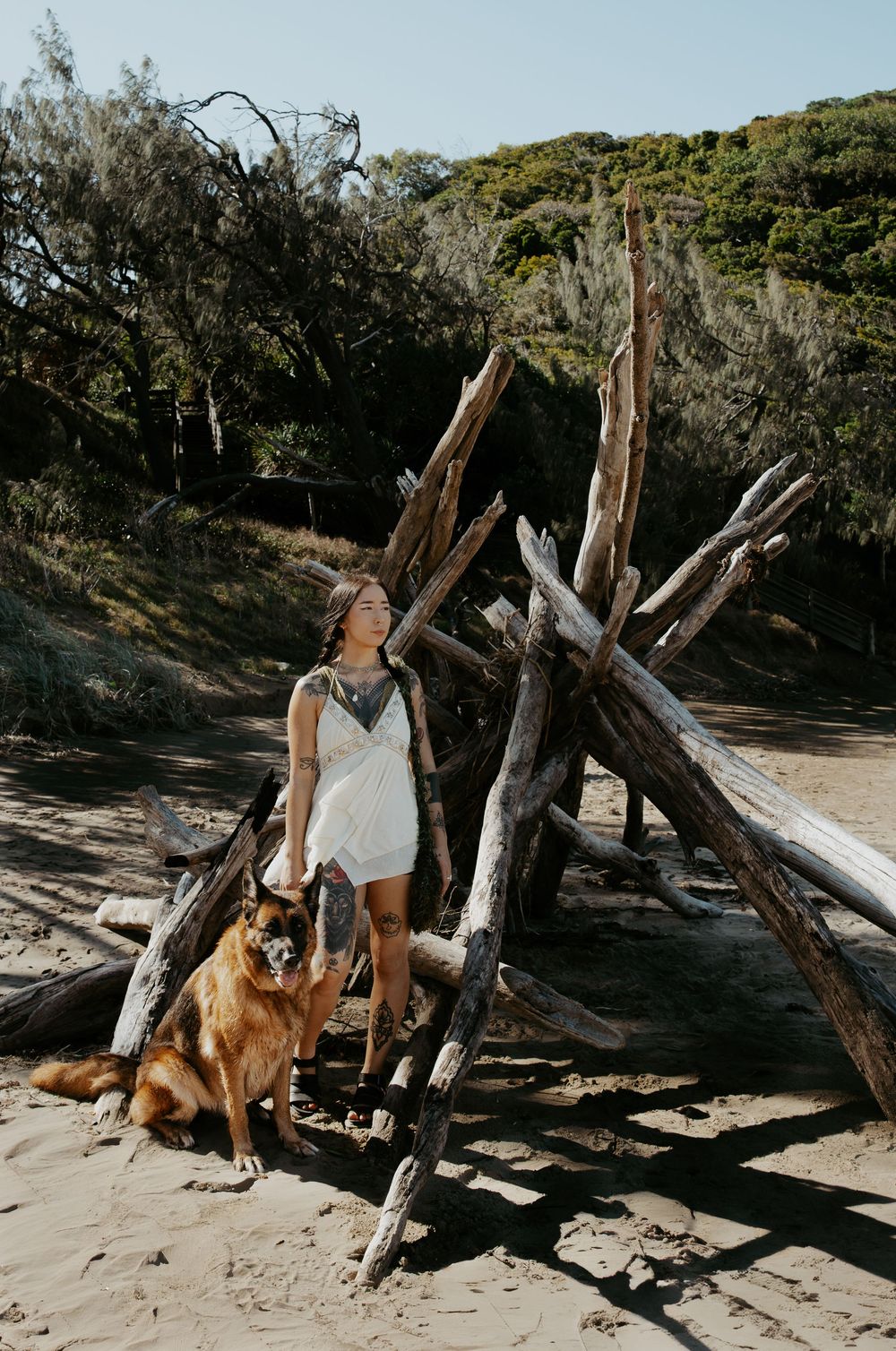 Woman in white dress stands with a brown dog beside a driftwood teepee on a sandy beach.