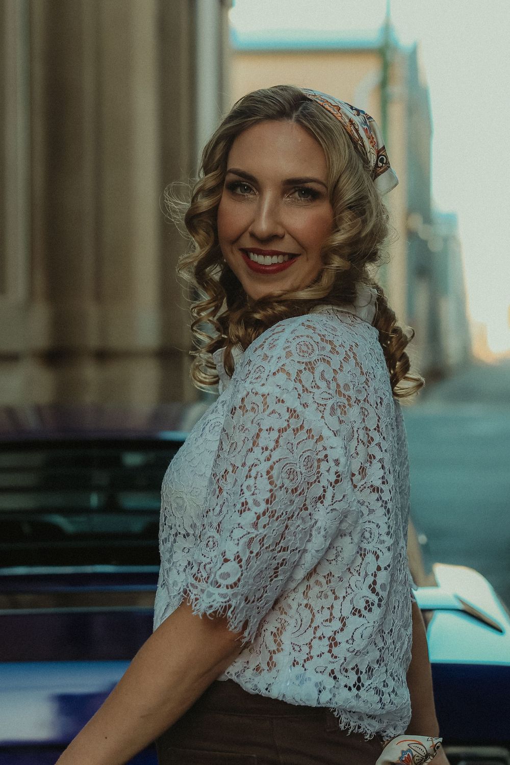 A smiling woman in a white lace top and headscarf poses beside a vintage car on a city street.