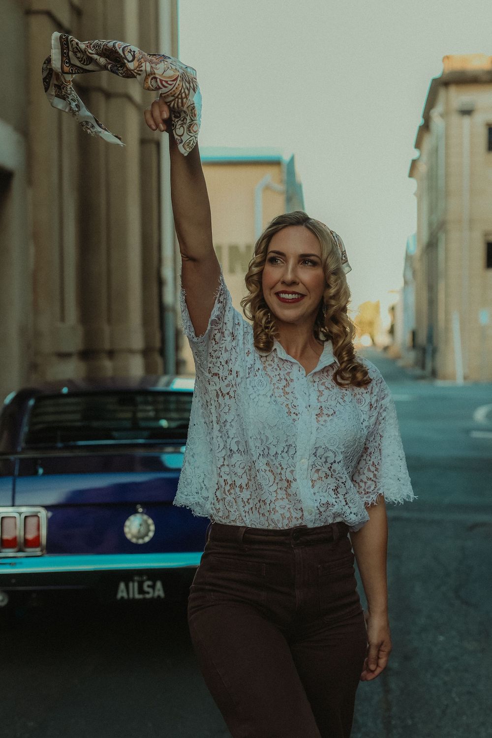 Woman in white lace top and brown pants waving a scarf on a city street with a vintage blue car behind her.