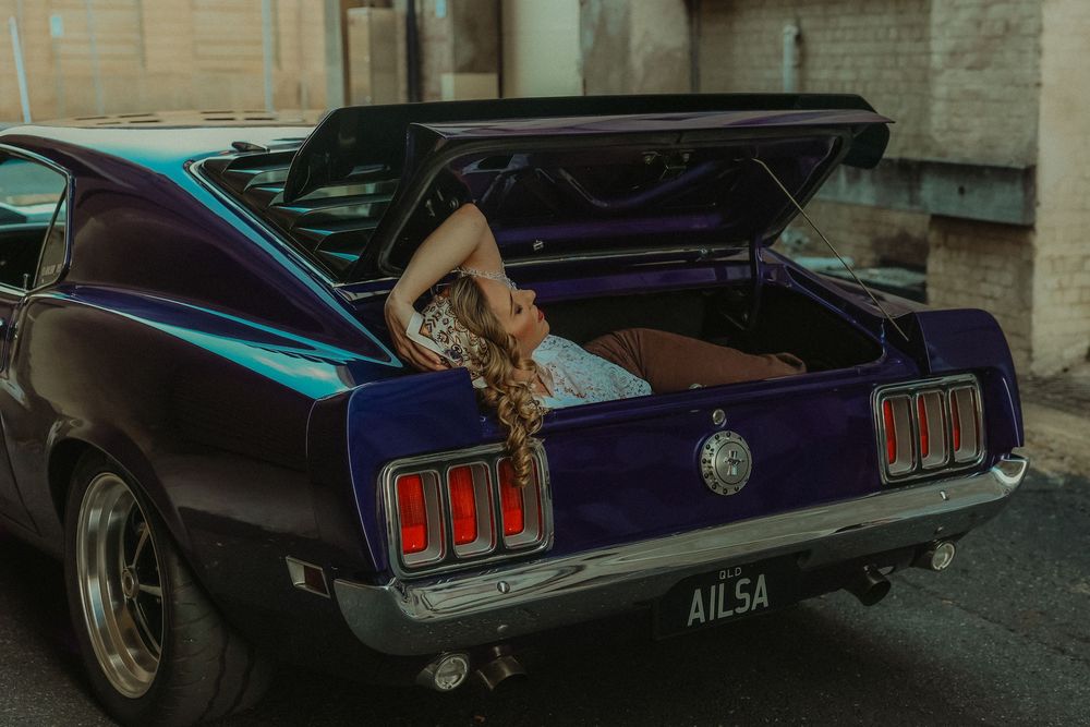 A woman with long blonde hair reclines in the open trunk of a classic purple Ford Mustang named Ailsa.