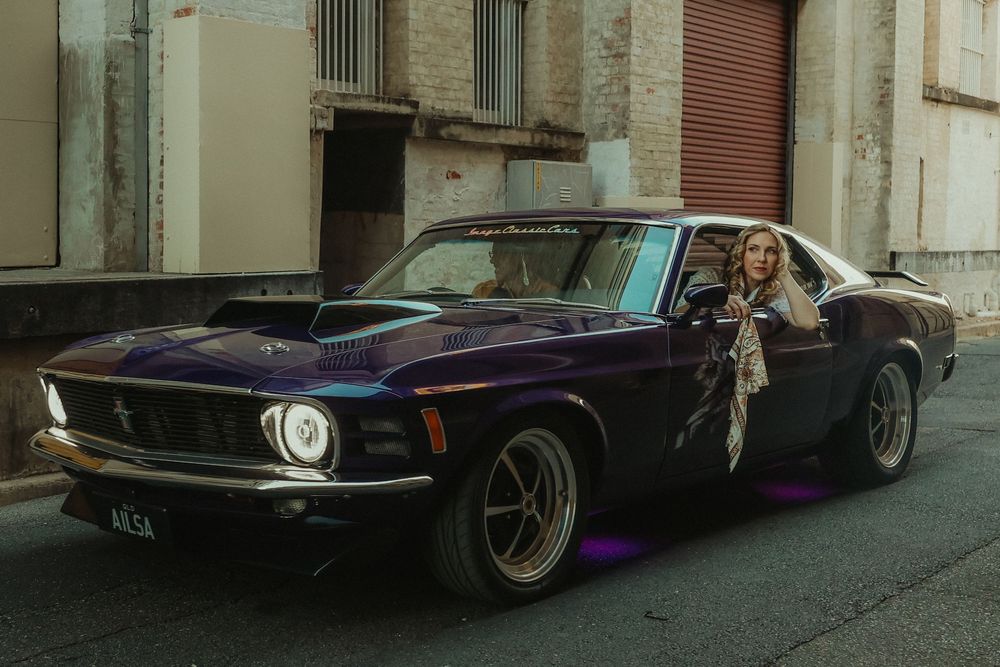 Woman leaning out of a classic purple Ford Mustang muscle car parked on a city street.