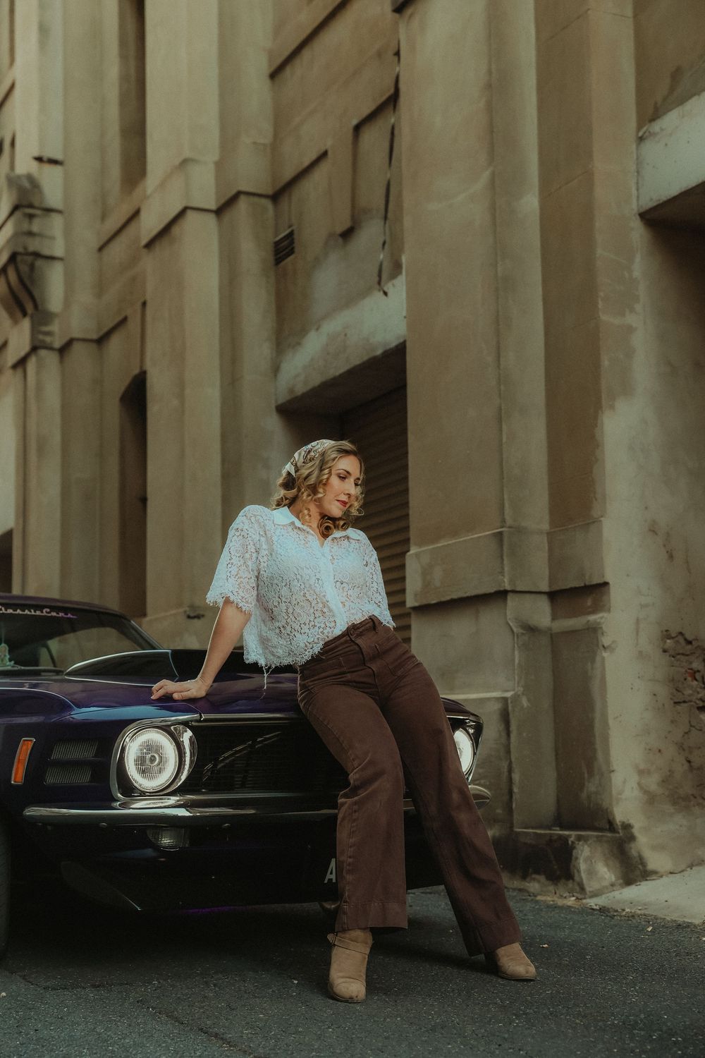 Woman in lace blouse and brown trousers leaning on vintage purple muscle car on urban street.