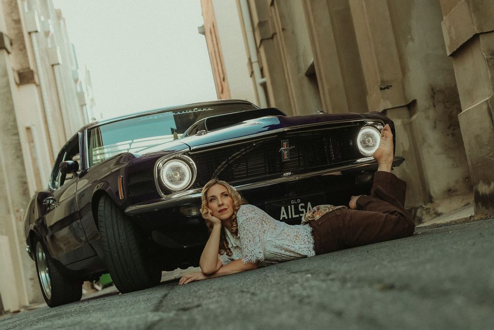 A blonde woman lies on the ground in front of a classic black Ford Mustang in a narrow urban alley.