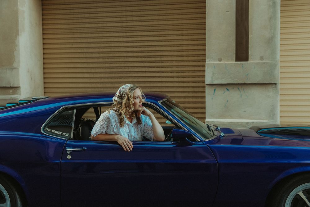 Smiling blonde woman in white lace dress leaning out of a classic blue muscle car near a building.