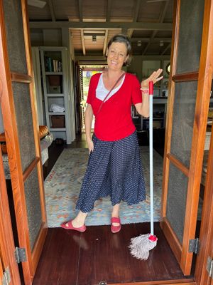 Smiling woman in red top and polka dot skirt holding a mop in a wooden doorway.