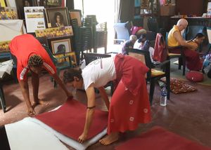 People rolling out a red carpet mat on the floor inside a room with chairs and decorative items in the background.