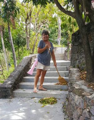 Woman sweeping outdoor stone steps with a broom surrounded by lush tropical trees and greenery.