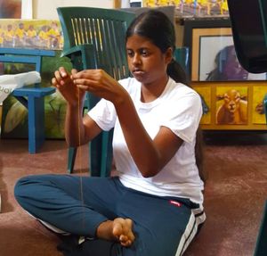 Young girl sitting cross-legged on floor, carefully threading a string with focused concentration indoors.