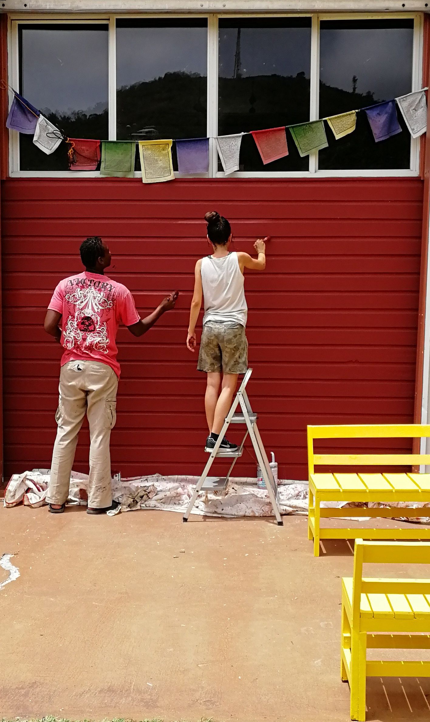 Two people painting a red wall outdoors, one standing on a ladder, with colorful prayer flags hanging above.