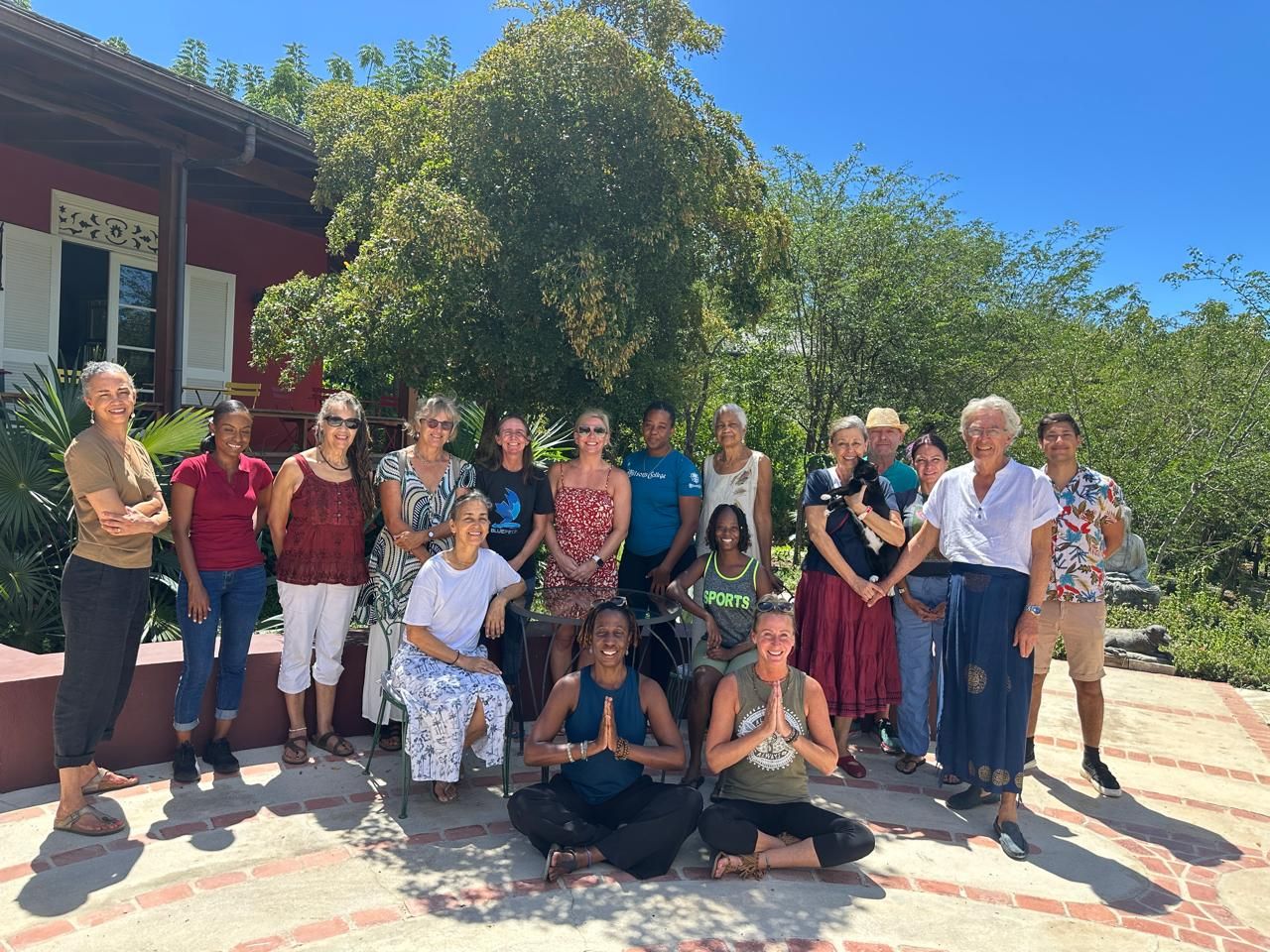 Group of diverse women posing outdoors at a yoga retreat, surrounded by lush greenery and a red building.