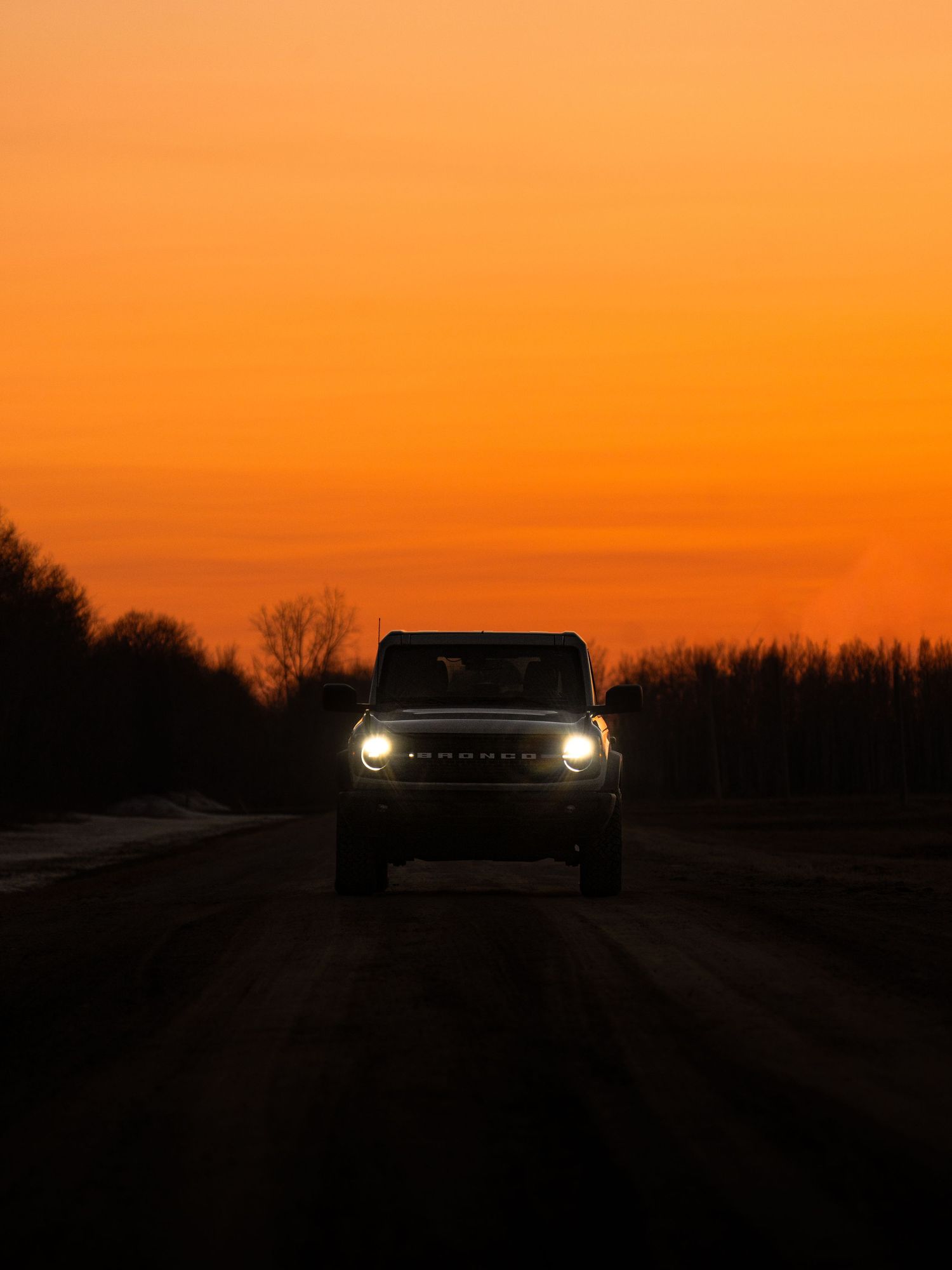 A Ford Bronco drives toward the camera on a rural road against a vivid orange sunset sky.
