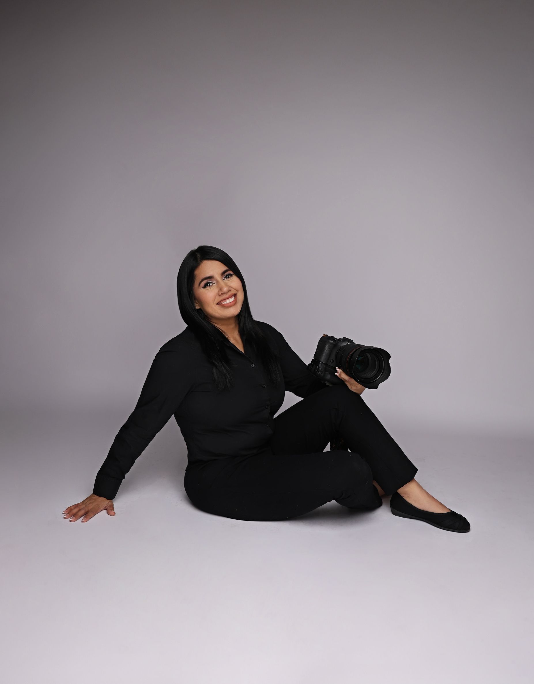 A smiling woman in all-black outfit sits on studio floor holding a professional camera, posing confidently.