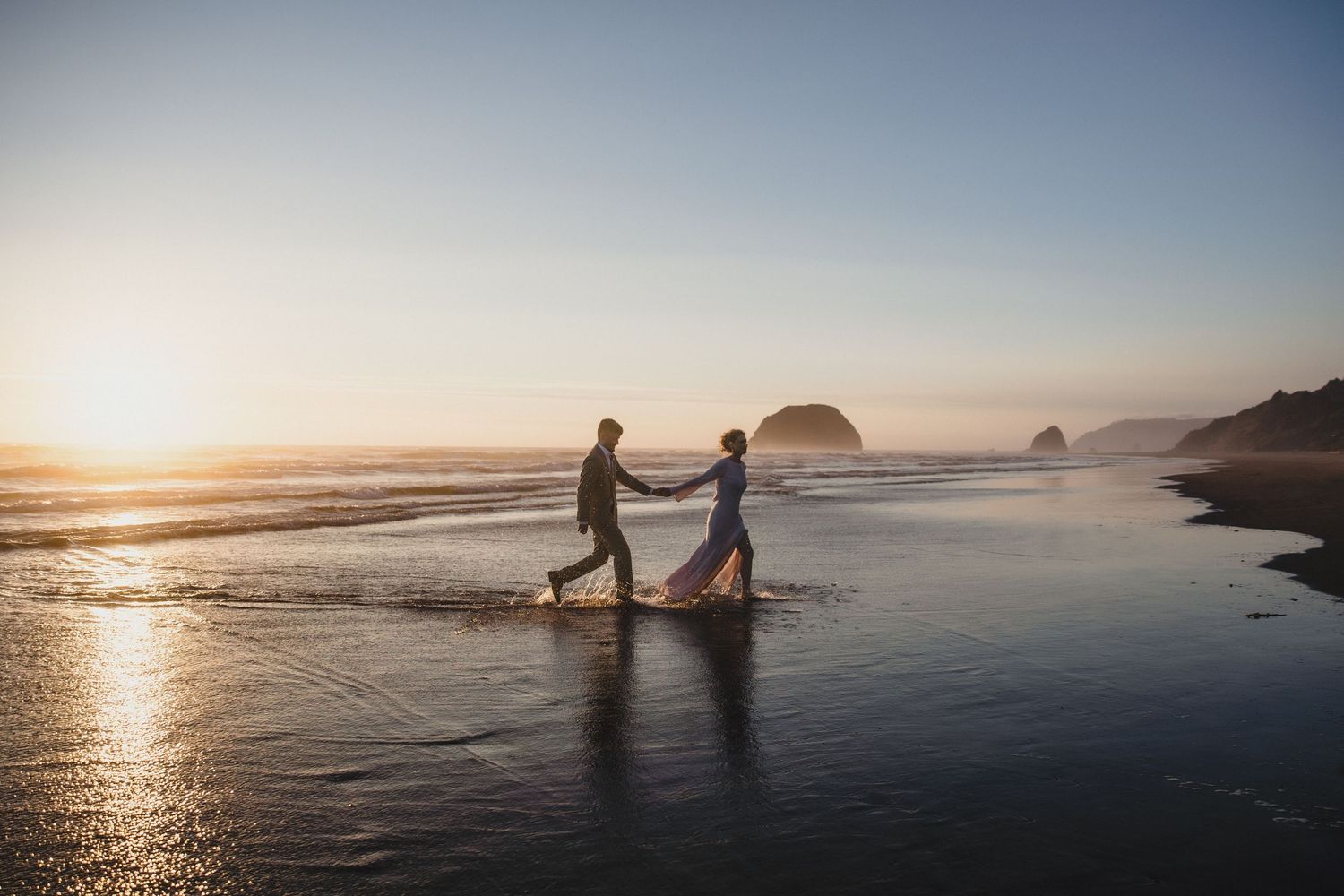 Couple holding hands walking on a sunlit beach at sunset with ocean waves and rocky silhouettes in the background.