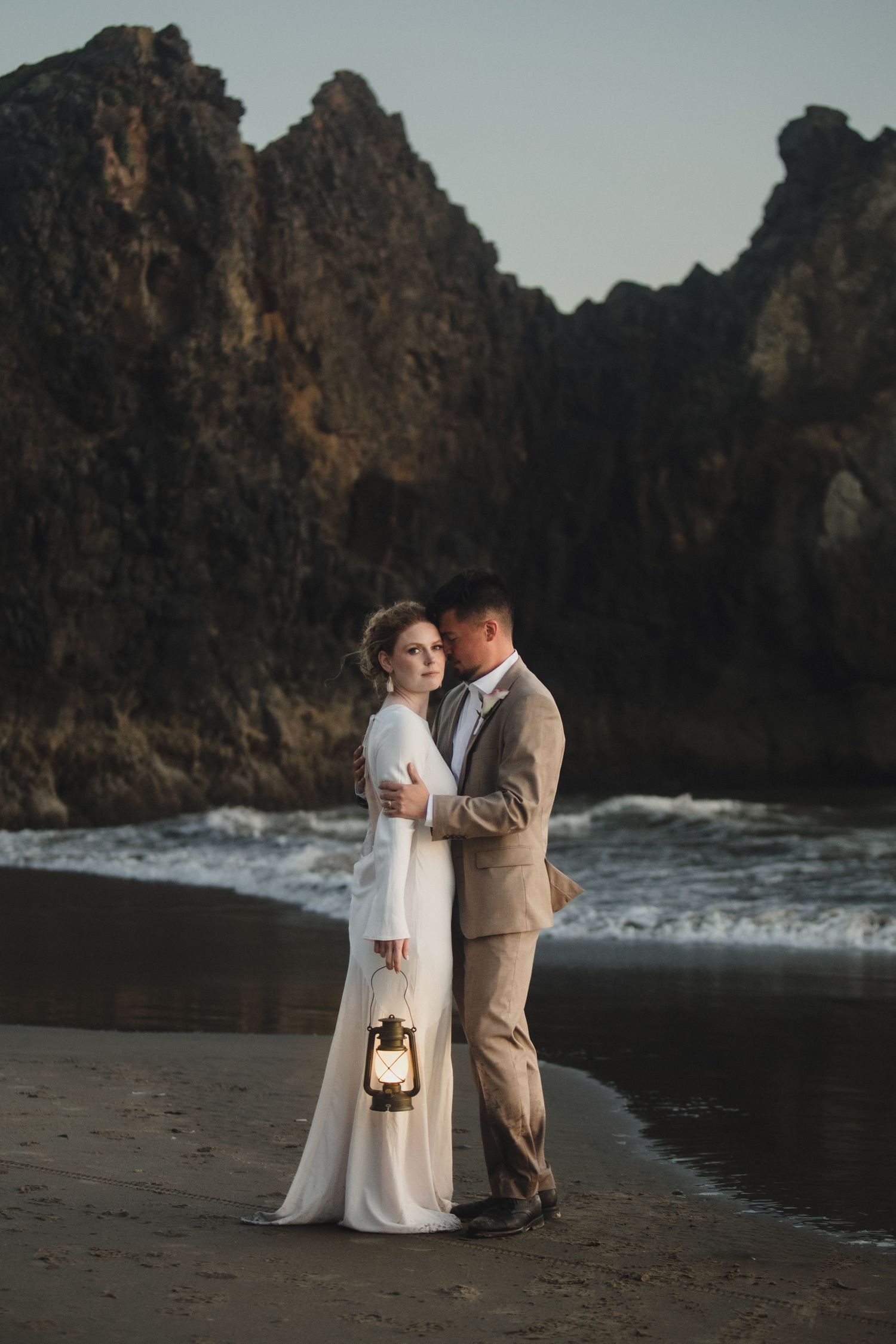 Bride and groom embrace on a dark beach at dusk, holding a vintage lantern with dramatic rocky cliffs behind them.