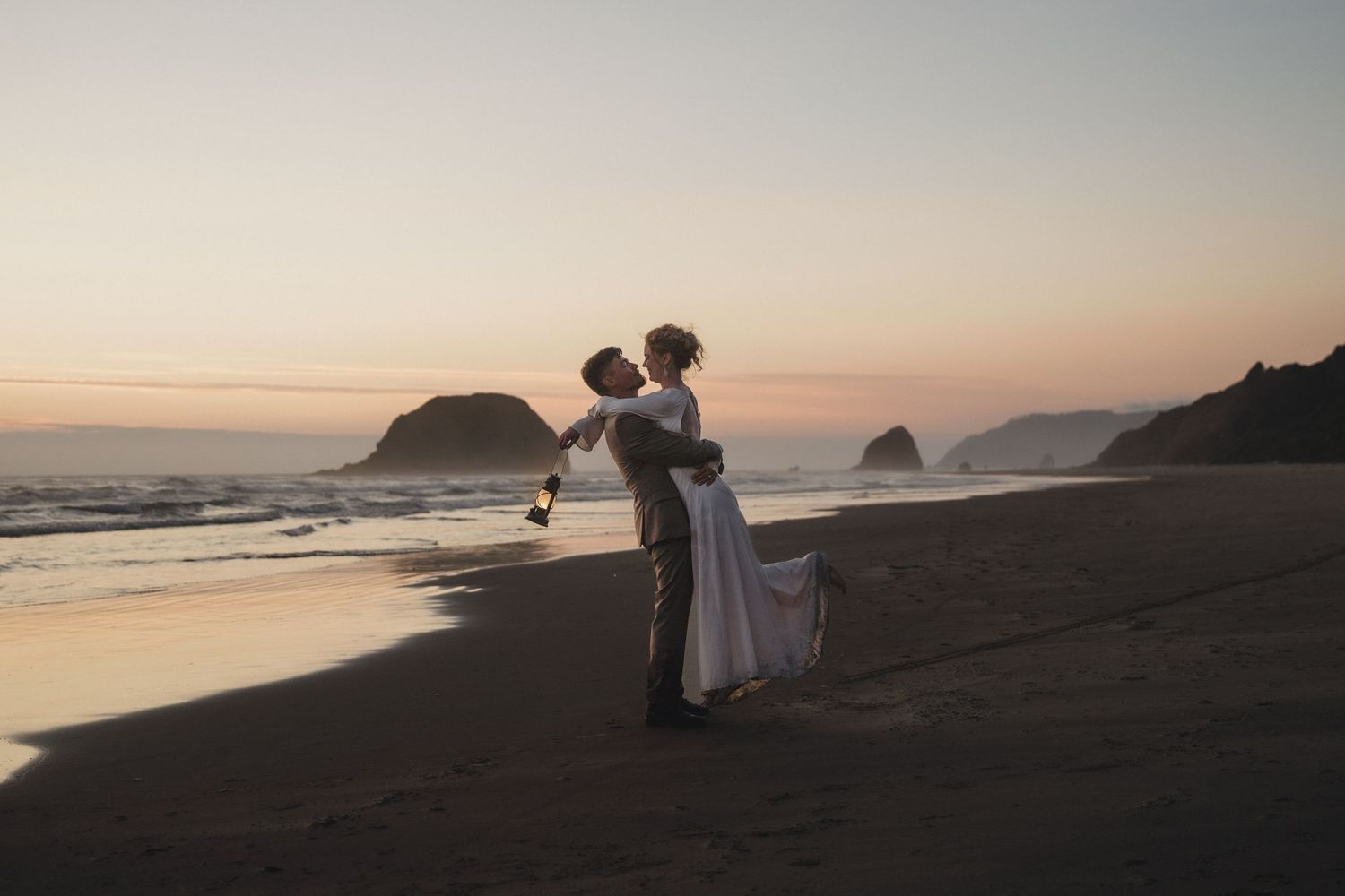Bride and groom share a romantic kiss on a sandy beach at sunset with ocean waves and rocky cliffs in the background.