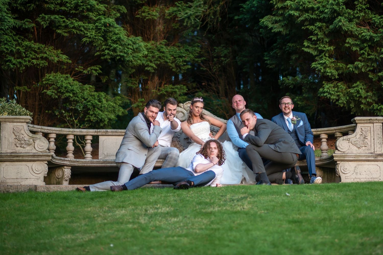 Wedding party posing playfully on green lawn with stone balustrade and lush trees at elegant outdoor venue.