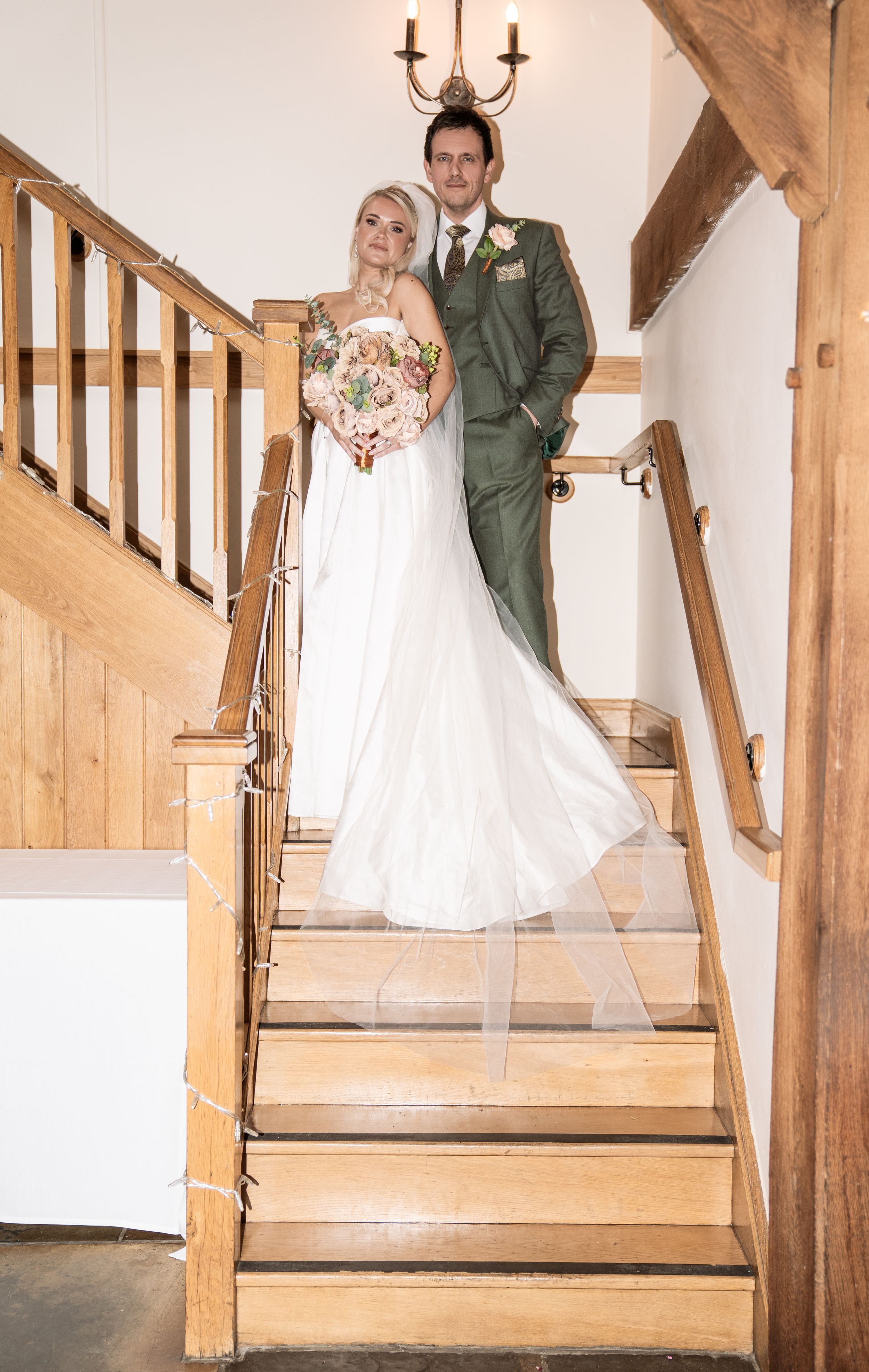 Bride and groom pose together on wooden staircase, bride holding pink bouquet in white gown, groom in green suit.