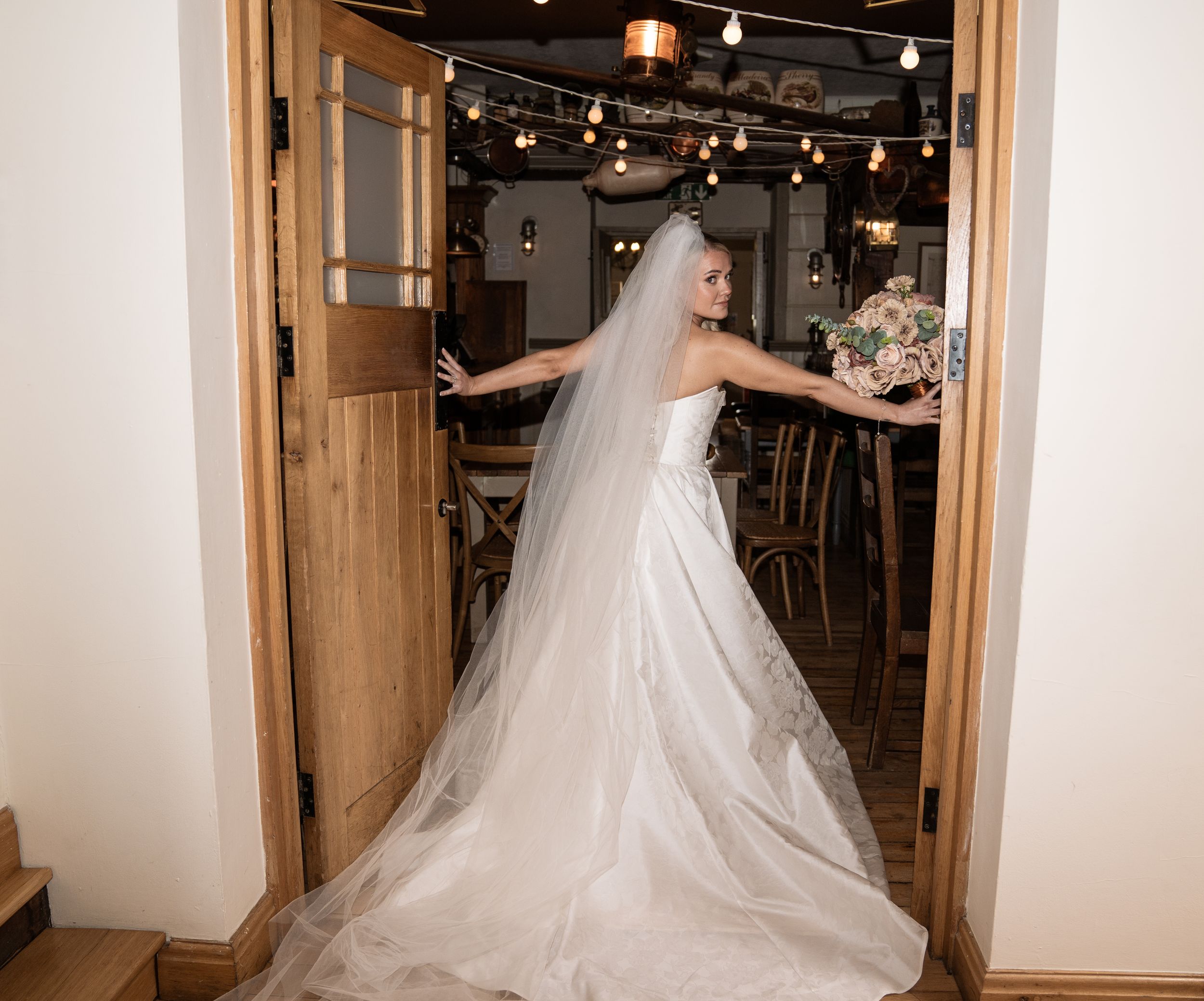 Bride in white gown and veil holds bouquet while standing in rustic wooden doorway of elegant wedding venue.