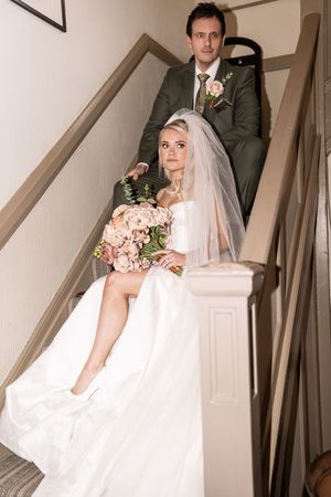 Bride seated on staircase holding pink floral bouquet with veil, groom standing behind in green suit.