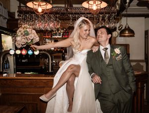 Bride and groom laughing together at a pub bar, bride holding floral bouquet in wedding dress and groom in green suit.