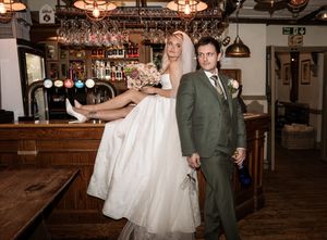 Bride sitting on a pub bar held by groom in green suit, wedding photo in rustic British pub setting.