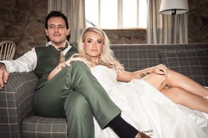 Elegant couple in wedding attire relaxing on a sofa, groom in green suit, bride in white dress with tattoo.