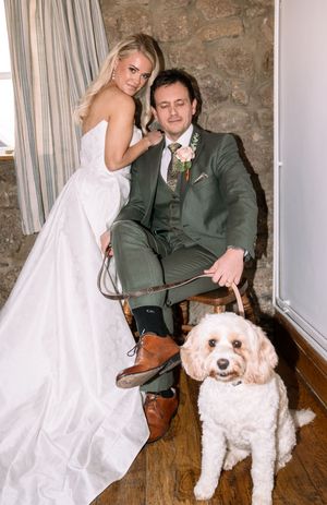 Bride in white gown and groom in green suit pose with a fluffy white Cavapoo dog in a rustic stone venue.