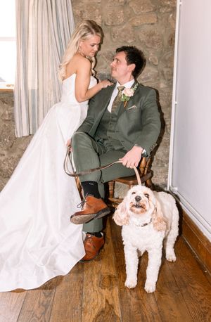 Bride in white gown and groom in green suit sit with their fluffy dog on a wooden floor at their wedding.