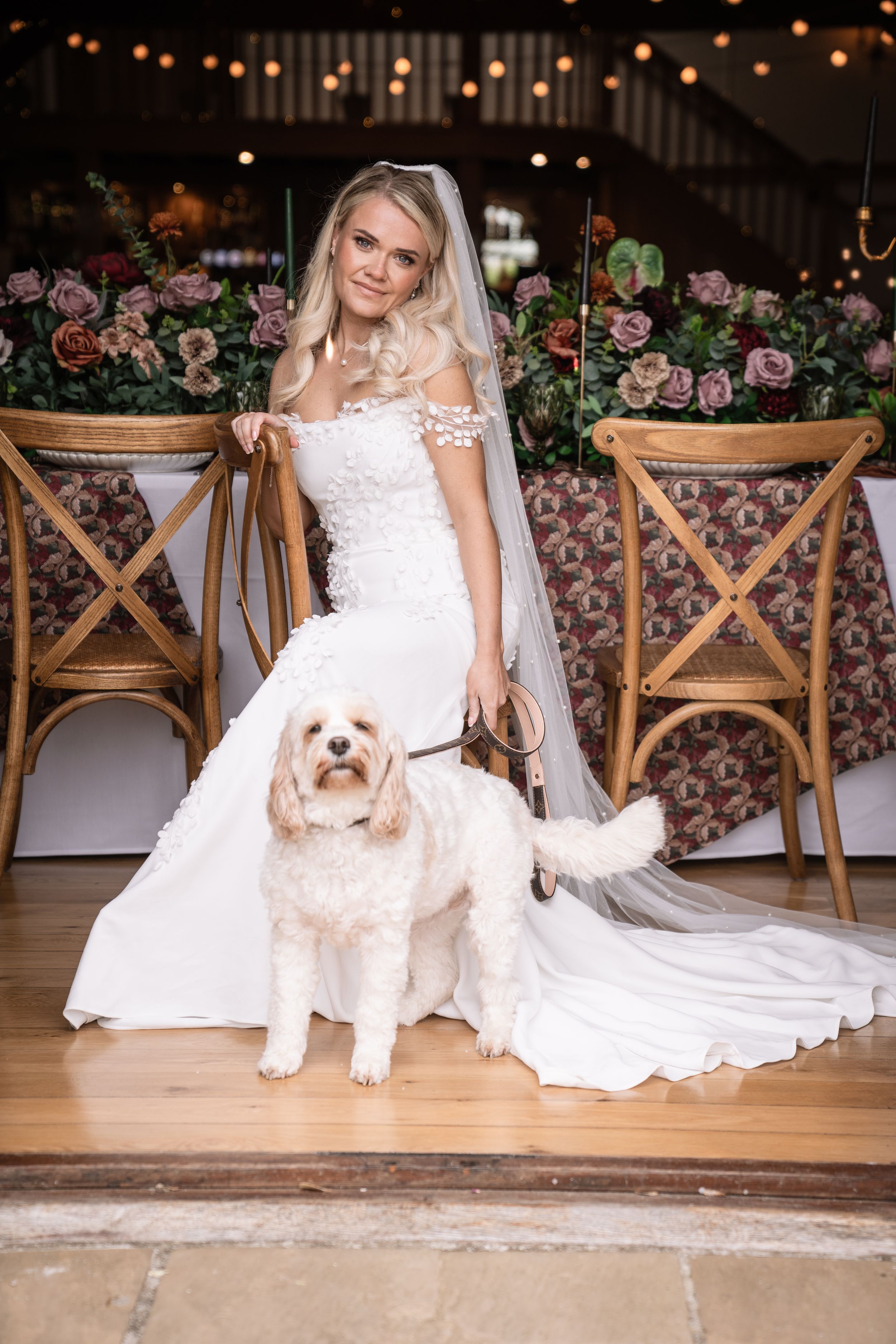 Blonde bride in lace wedding dress and veil poses with a white Cavapoo dog in a floral decorated barn venue.