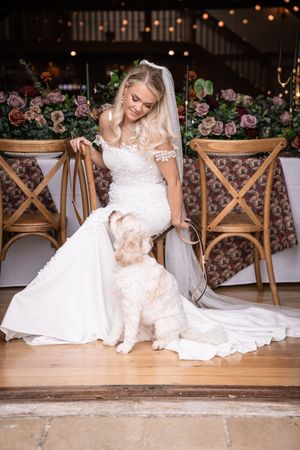 Blonde bride in lace wedding dress and veil posing with fluffy dog at wedding venue.
