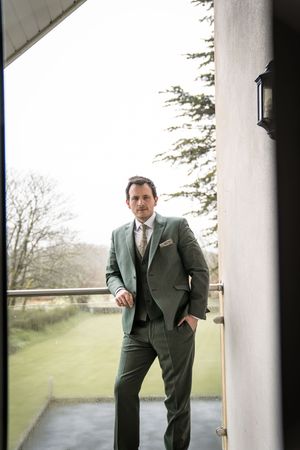 Groom in elegant green three-piece suit standing on outdoor balcony with garden views in background.