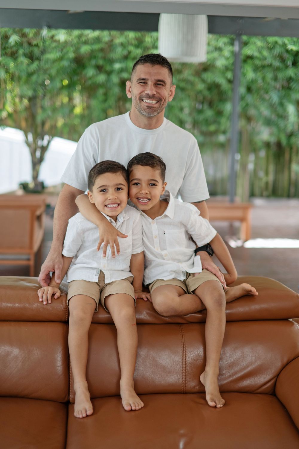 A smiling father in a white t-shirt poses with his two young sons on a brown leather sofa in a bright living room.